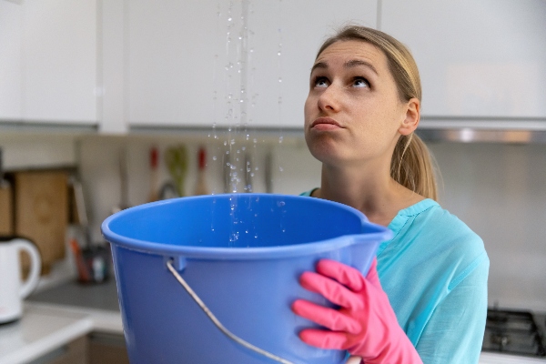 A frustrated homeowner holds up a bucket to catch water leaking from the ceiling.
